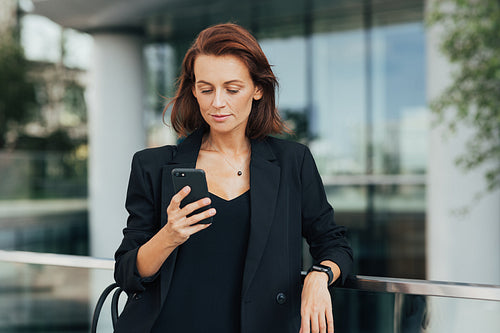 Middle-aged businesswoman in black formal wear looking at her smartphone while standing outdoors