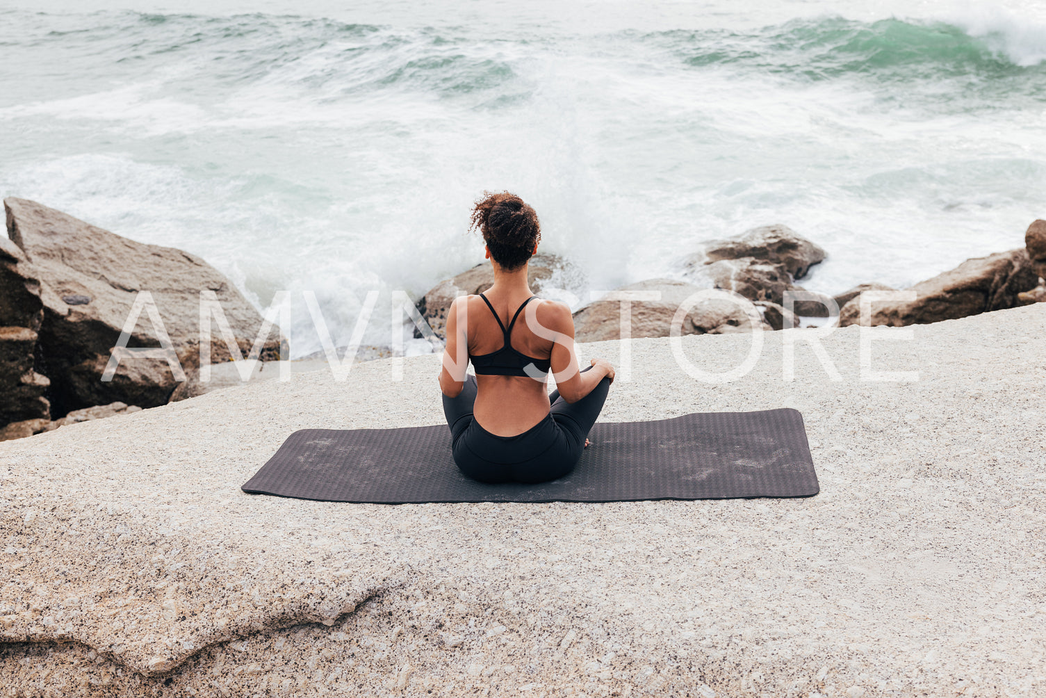 Rear view of a woman sitting on a mat in lotus pose and looking at waves and splashes