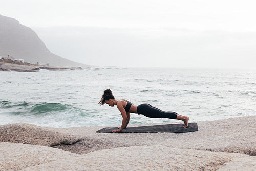 Side view of young woman in plank pose exercising on mat by ocean at evening