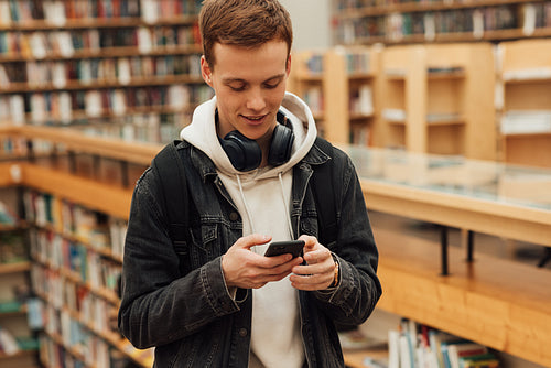 Young smiling guy with smartphone standing in library