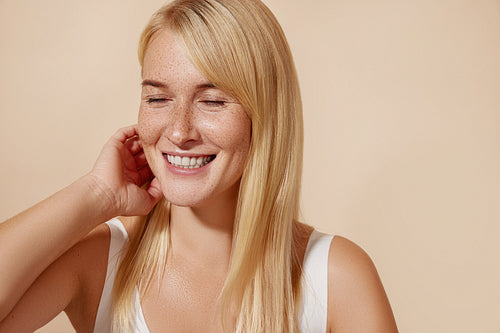 Cheerful woman with closed eyes adjusting her blond hair. Happy female with freckles against a pastel backdrop.