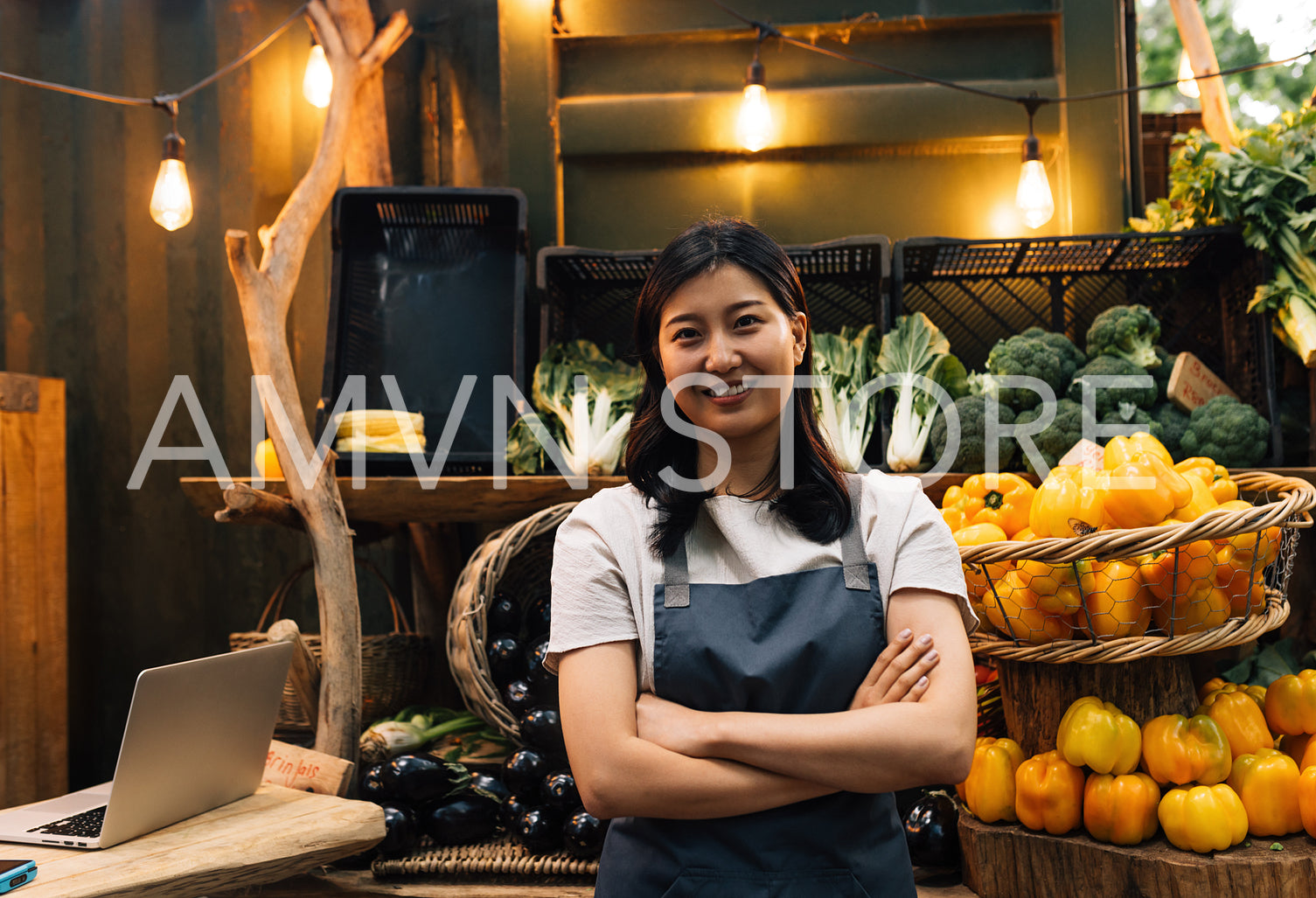 Portrait of an outdoor market owner. Asian woman with crossed arms wearing an apron standing against a stall.