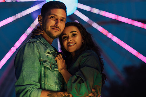 Young lovely couple hugging at night standing against the Ferris wheel. Girlfriend and boyfriend are standing together in an amusement park during the festival.