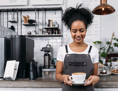 Smiling barista in an apron looking at a cup of coffee. Woman with curly hair works as a barista in a coffee shop.