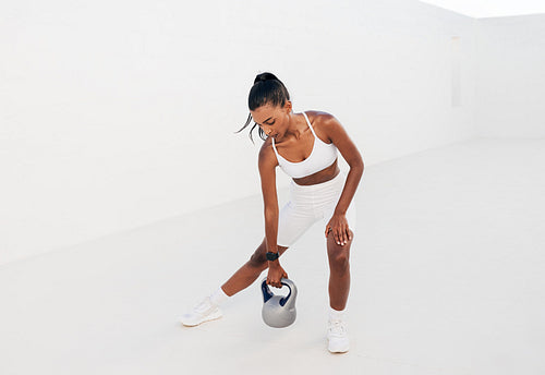 Slim female in a white sports bra doing intense training with a kettlebell. Full length of a fitness influencer outdoors.