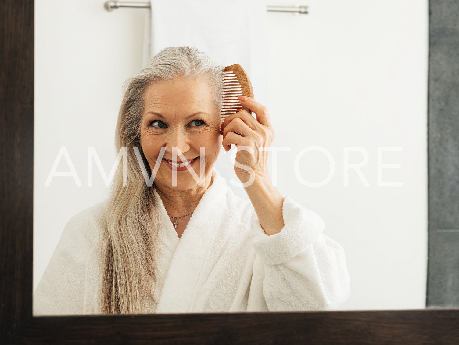 Woman holding a wood comb in the bathroom. Aged female combing her hair while looking at a mirror.