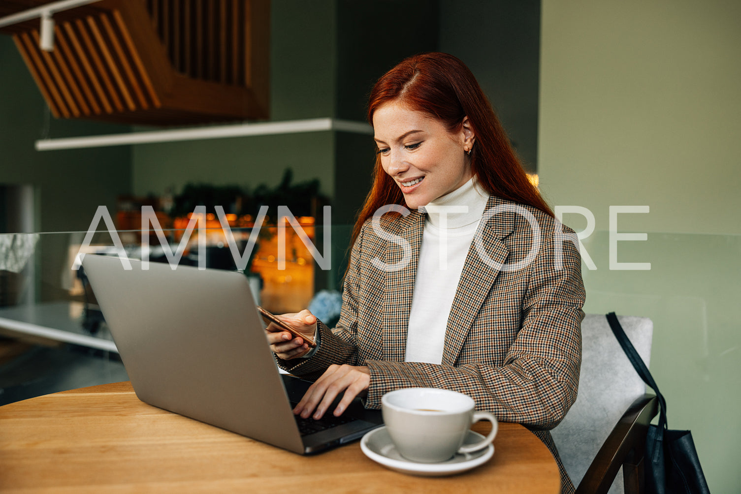 Smiling redhead woman typing on laptop and holding mobile phone while sitting in a cafe