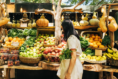 Side view of a young woman with a bag looking at vegetables and fruits at a local outdoor market.