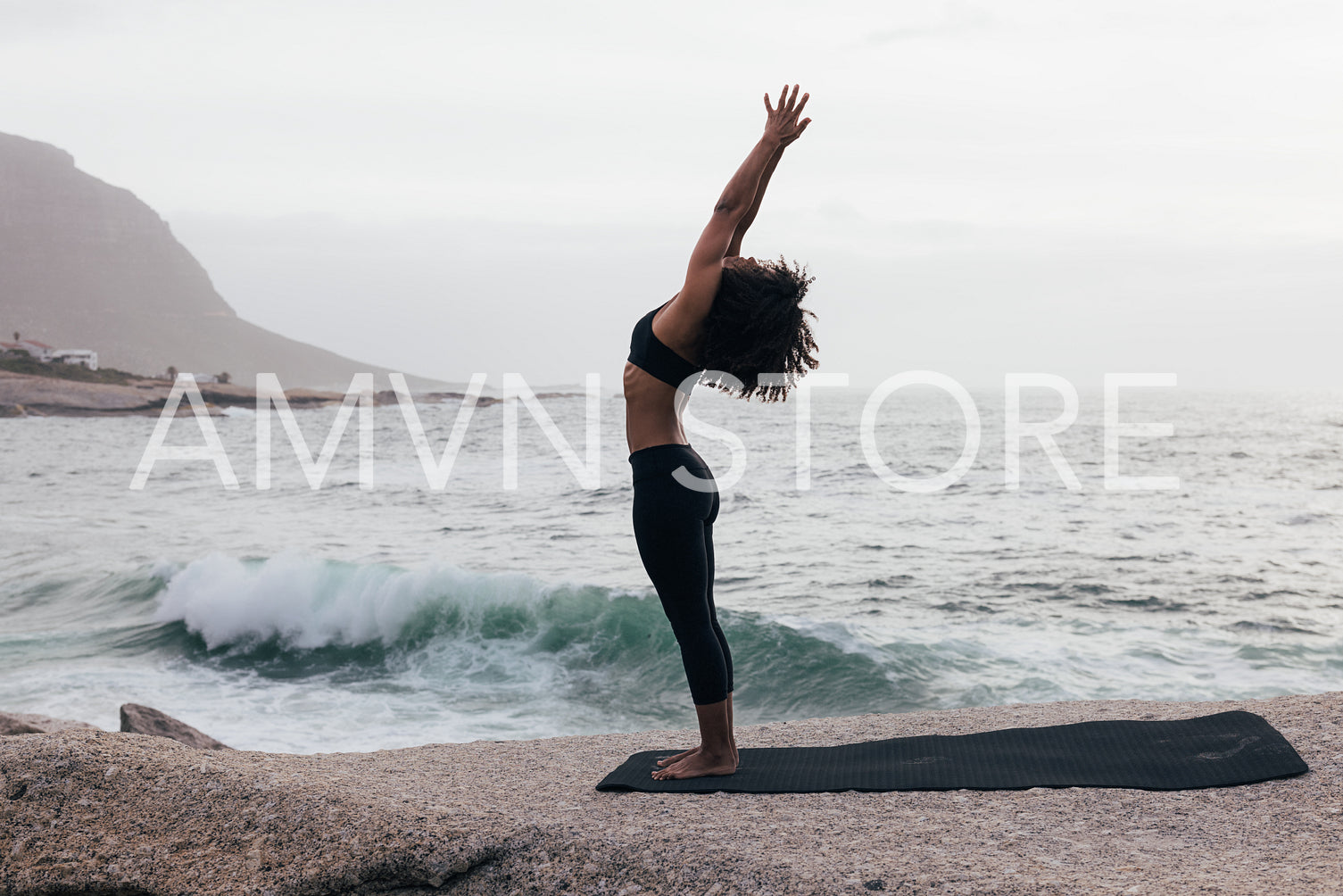 Side view of a slim female raising hands in yoga pose standing against an ocean at evening