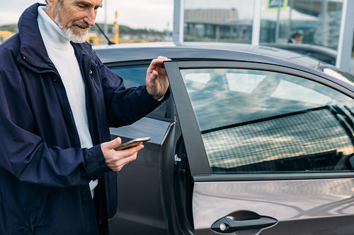 Mature man opening a door of his car. Bearded man looking on smartphone outdoors.