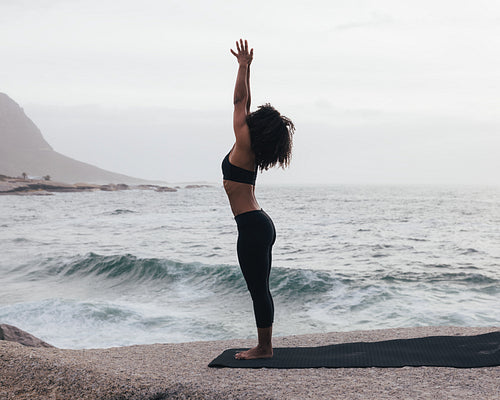 Woman practicing Urdhva Hastasana pose by ocean. Slim female standing outdoors in Raised Hands Pose.