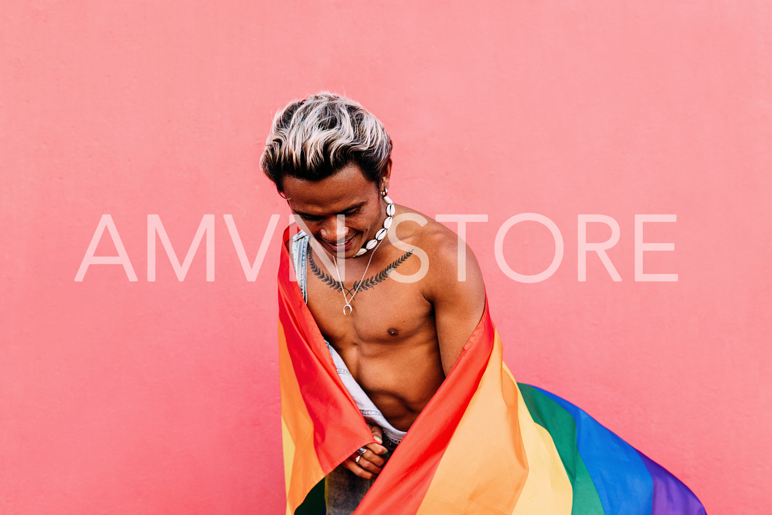 Young bare chested guy having fun while standing against pink wall with LGBT flag