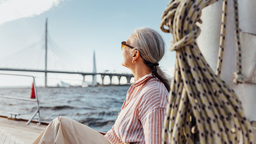 Senior woman with gray hair wearing sunglasses looking into the distance while sitting on a sailboat