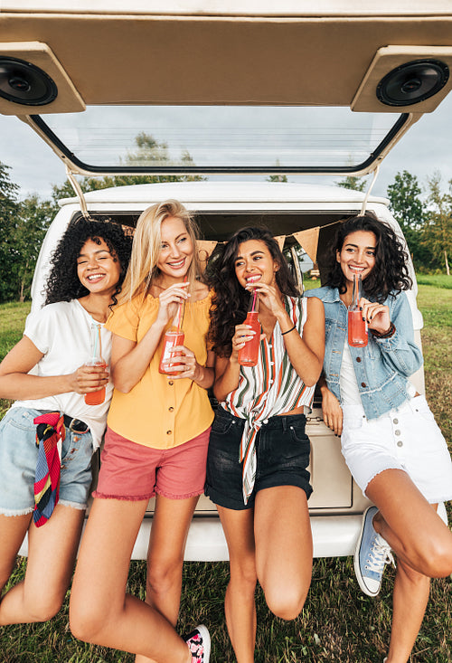 Group of diverse women holding cocktails while standing at the back of camper van. Four females with bottles standing at a car during summer vacation.