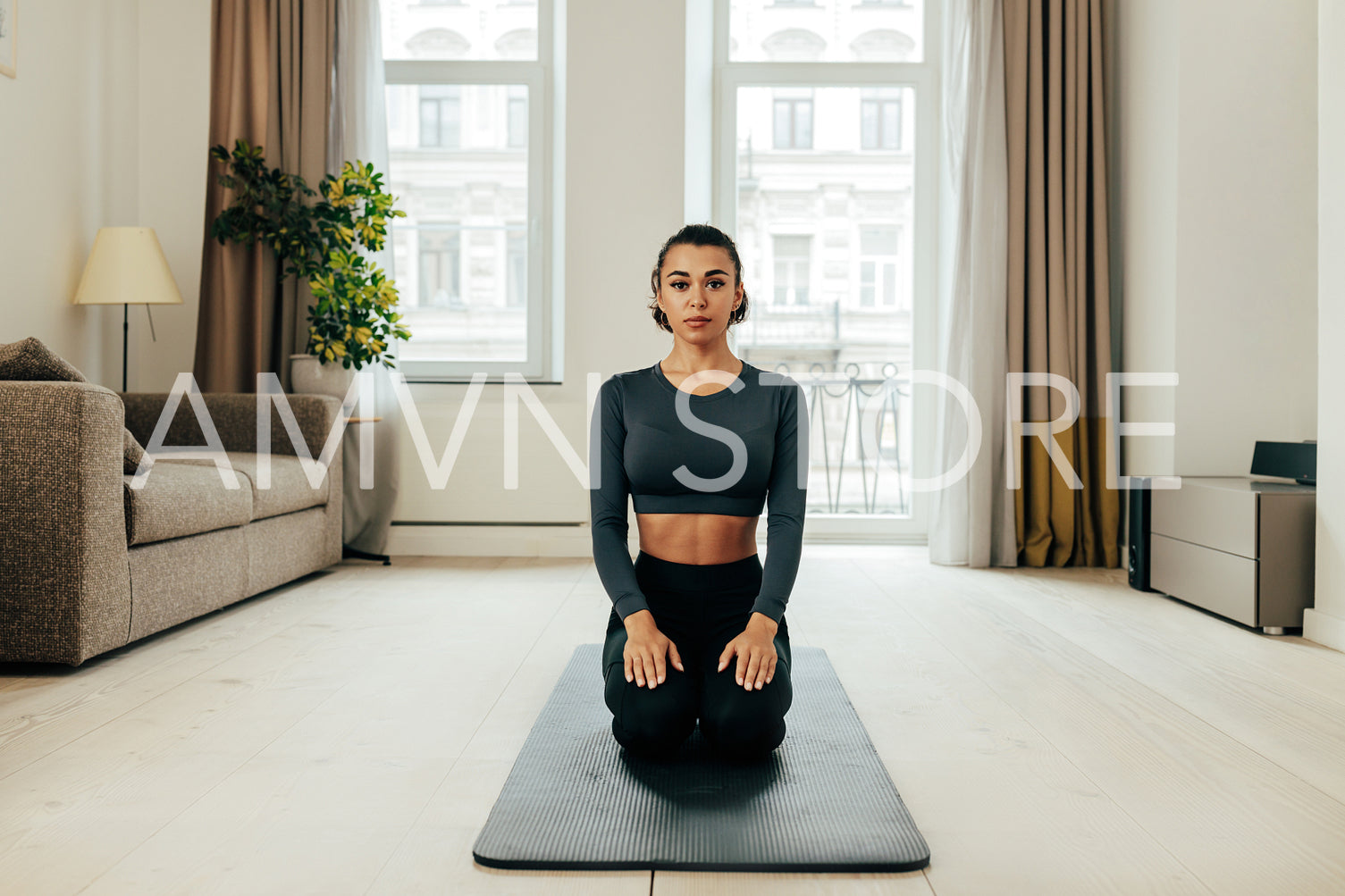 Young woman sitting on a mat in a yoga pose in living room and looking straight of a camera	