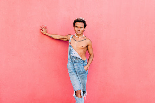 Young handsome man wearing jeans overall leaning pink wall looking at camera