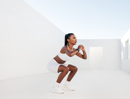 Female fitness influencer doing sit-ups in a white outdoor studio. Young woman doing workout outdoors. Female in white fitness attire is doing exercises for her glutes.