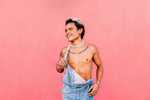 Portrait of a young smiling guy in denim overalls looking away while standing at pink wall