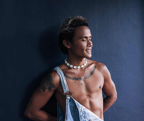 Smiling guy with closed eyes leaning against a blue wall. Stylish male wearing overall with bare chest relaxing outdoors.