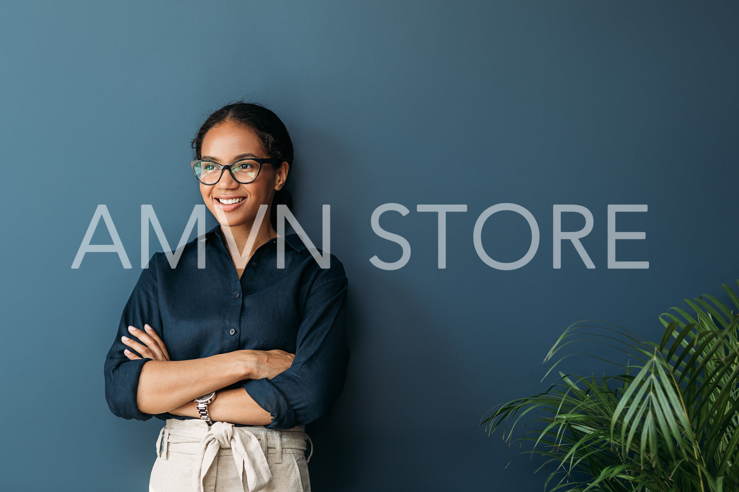 Confident businesswoman with crossed arms leaning on blue wall. Smiling entrepreneur in eyeglasses looking away.