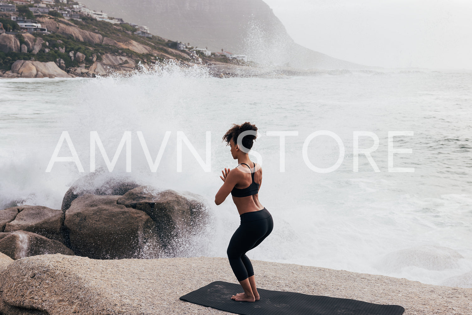 Woman practicing yoga while standing on stone against big waves and splashes. Female meditating at oceanside.