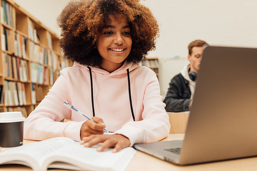 Smiling girl writing in a book while sitting at desk with laptop. Female student studying at college library.