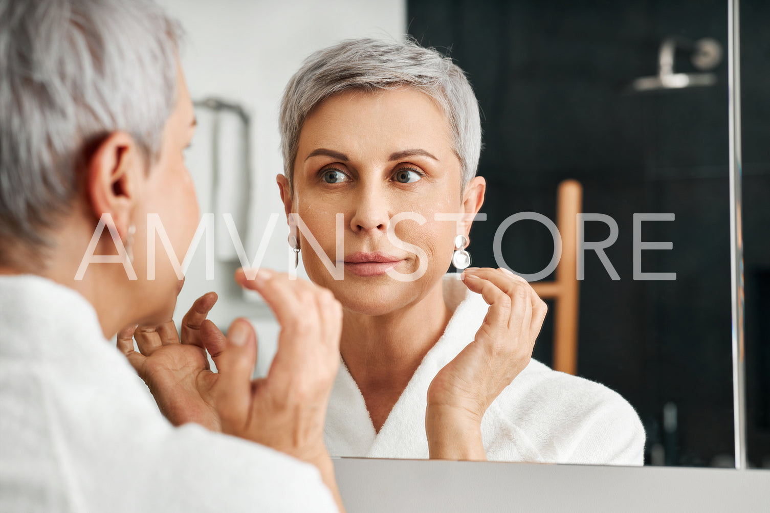 Smiling mature woman looking at her reflection while applying moisturizer on the face