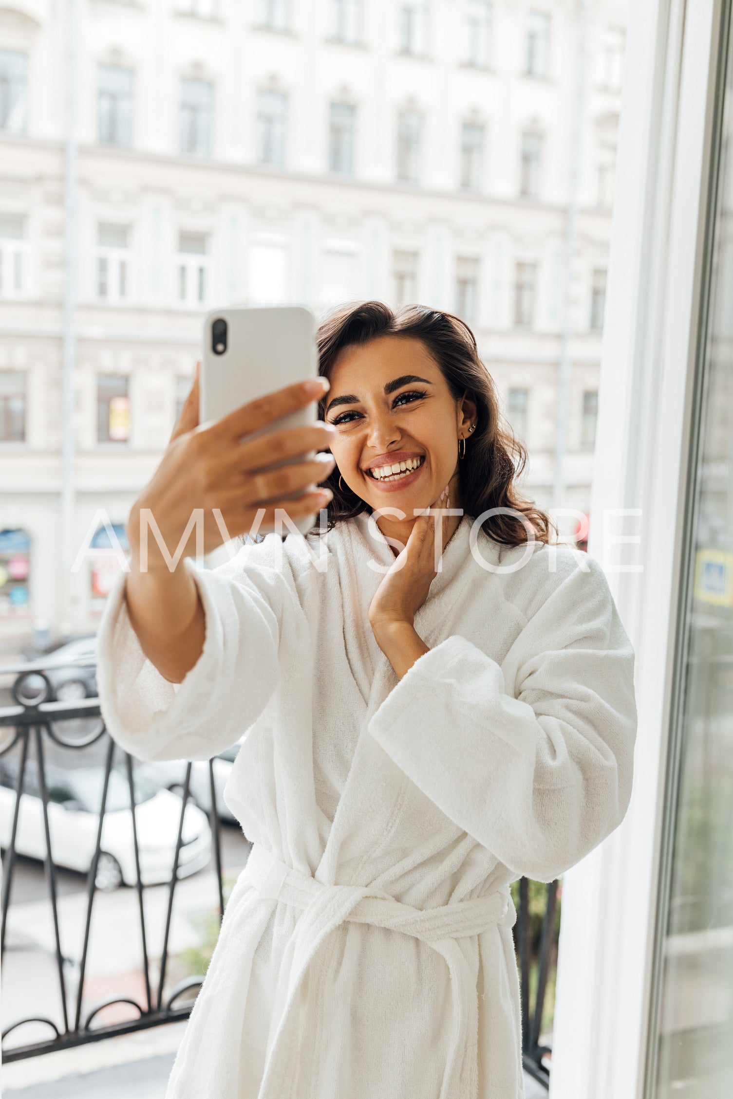 Happy woman taking selfie on a balcony. Young female in bathrobe holding a smartphone and making photos.