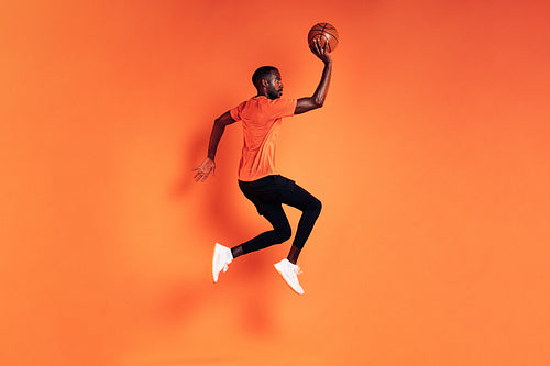 Male athlete jumping with basket ball. Side view of sportsman exercising over orange background.