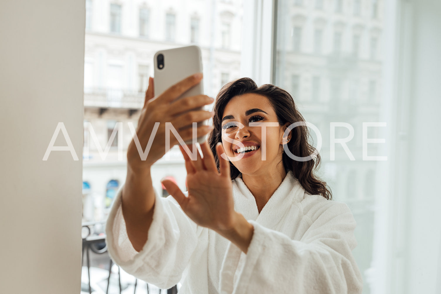 Happy beautiful woman taking a selfie on a balcony in her hotel room. Smiling female in bathrobe holding smartphone and making photographs on frontal camera.	