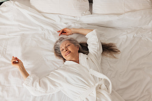 Happy senior woman in bathrobe lying on a bed in hotel. Cheerful aged female enjoying morning.