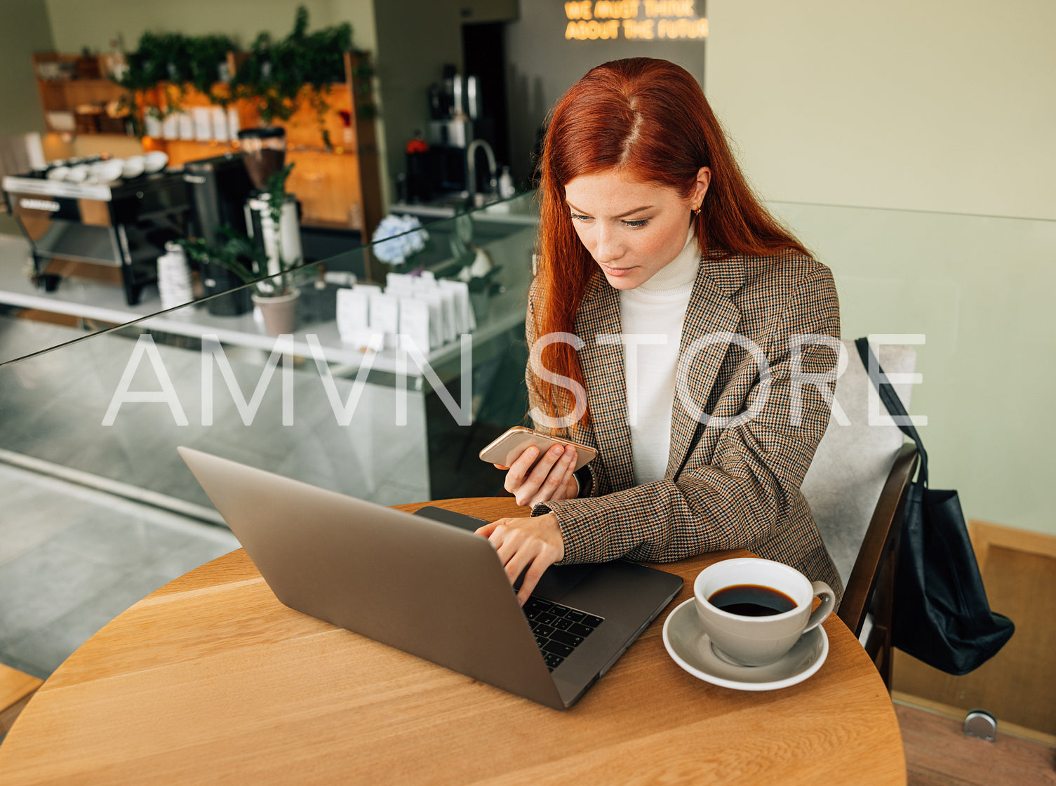 Young female entrepreneur working in a coffee shop. Woman with ginger hair typing on a laptop and holding mobile phone.