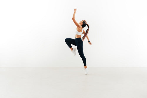 Side view of young female athlete jumping in the air raising hand up against white wall