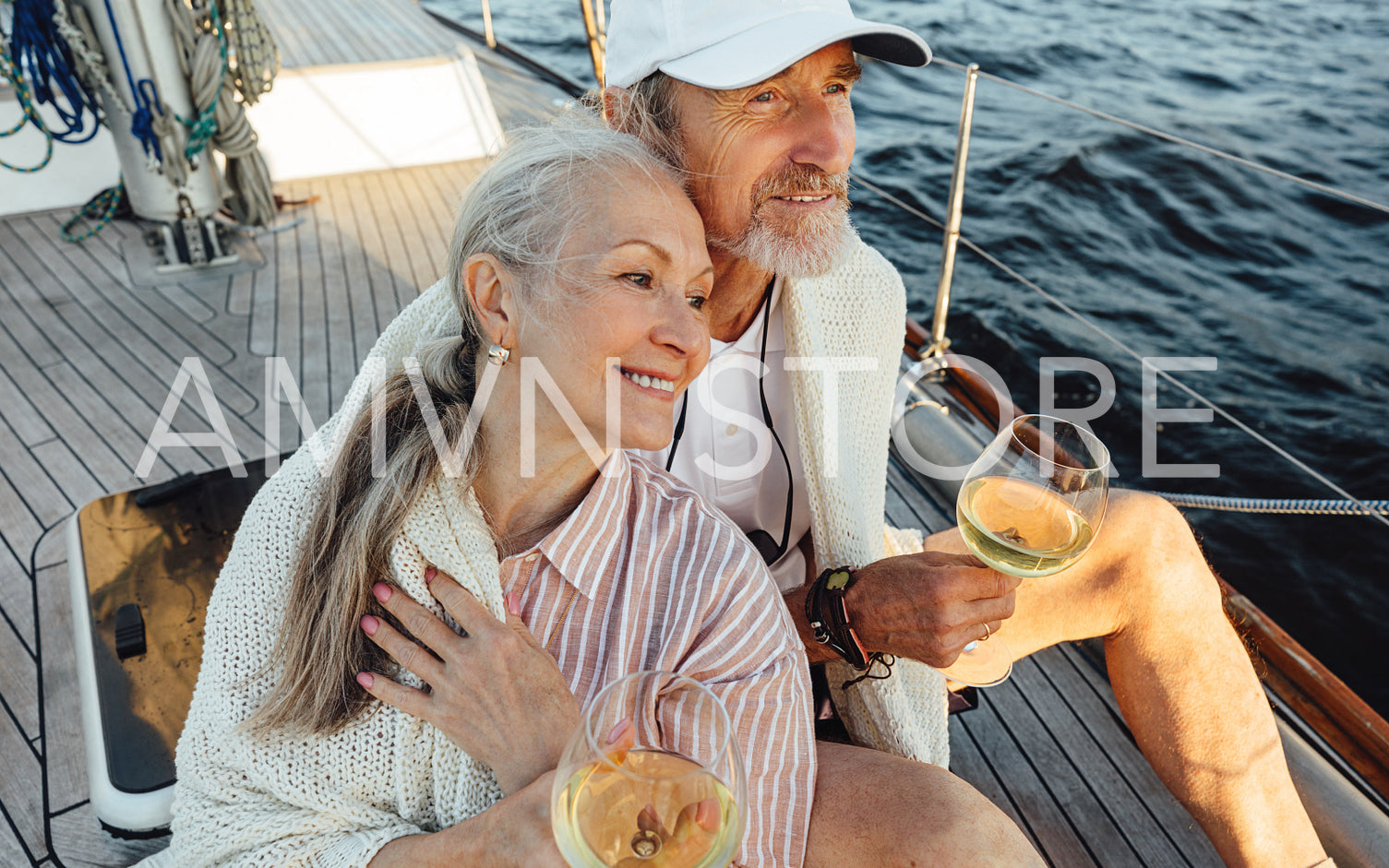 Senior couple wrapped with plaid and sitting on a yacht deck with bocals of wine. Two loving mature people sitting together on a sailboat and looking away.	