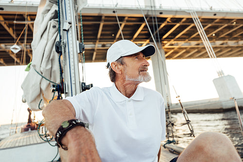 Relaxed mature man sitting on his yacht. Senior male in cap looking away while floating under a highway on a sailboat.