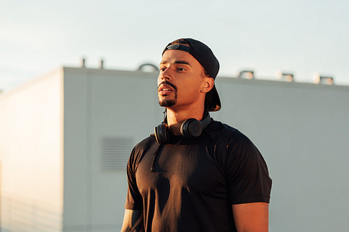 Portrait of a young confident male athlete in a cap. Athlete with wireless headphones standing on a rooftop at sunset.