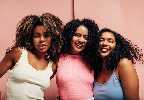 Three girlfriends in bright casual hugging each other looking at camera against a pink wall