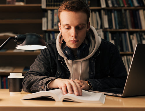 Male student reading from a book. Guy with headphones preparing his assignment while sitting in the evening in library.