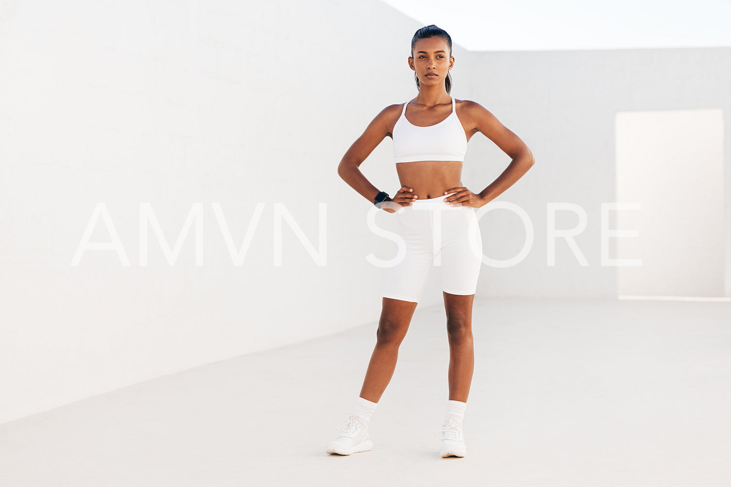 Portrait of a slim female in white fitness attire standing in an outdoor studio. Sportswoman relaxing with hands on hips.