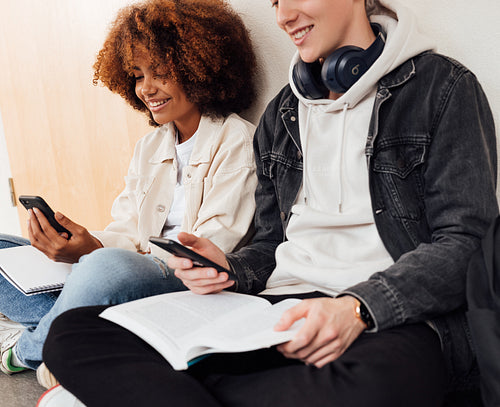 Smiling friends sitting together at wall with smartphones in school