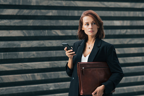 Confident businesswoman with ginger hair. Middle-aged woman holding smartphone and looking away. Female in formal clothes holding leather folder outdoors.