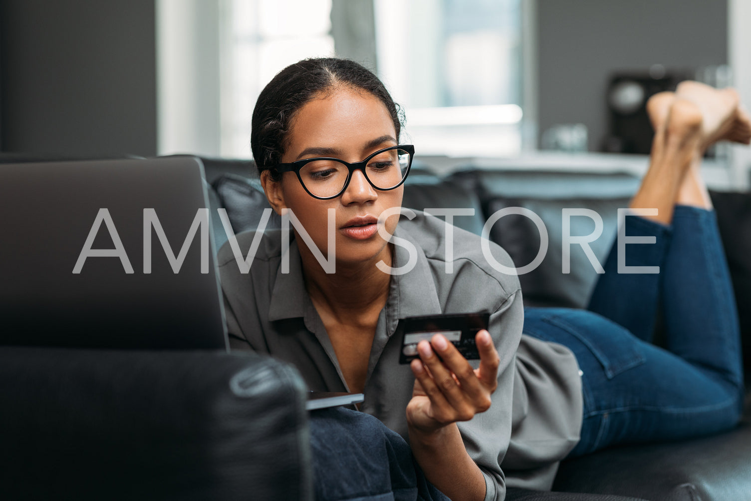 Young female in eyeglasses looking at a credit card while lying on a couch at home