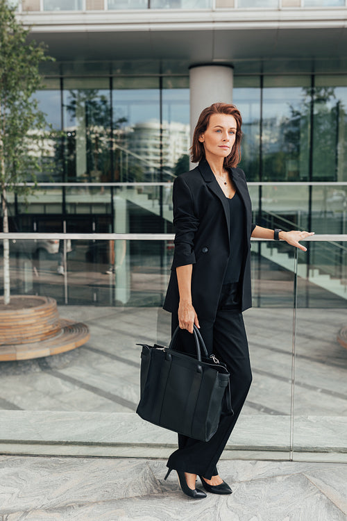 Full length of a middle-aged female in black formal wear leaning a railing standing against office building