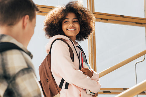 Smiling girl with books and backpack looking at classmate while walking on stairs