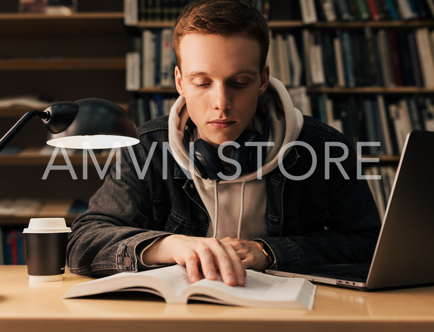 Male student reading from a book. Guy with headphones preparing his assignment while sitting in the evening in library.