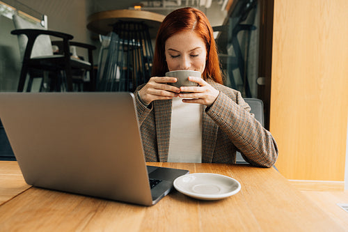 Smiling woman with ginger hair enjoying her morning coffee. Businesswoman holding a cup with closed eyes while sitting at a table.