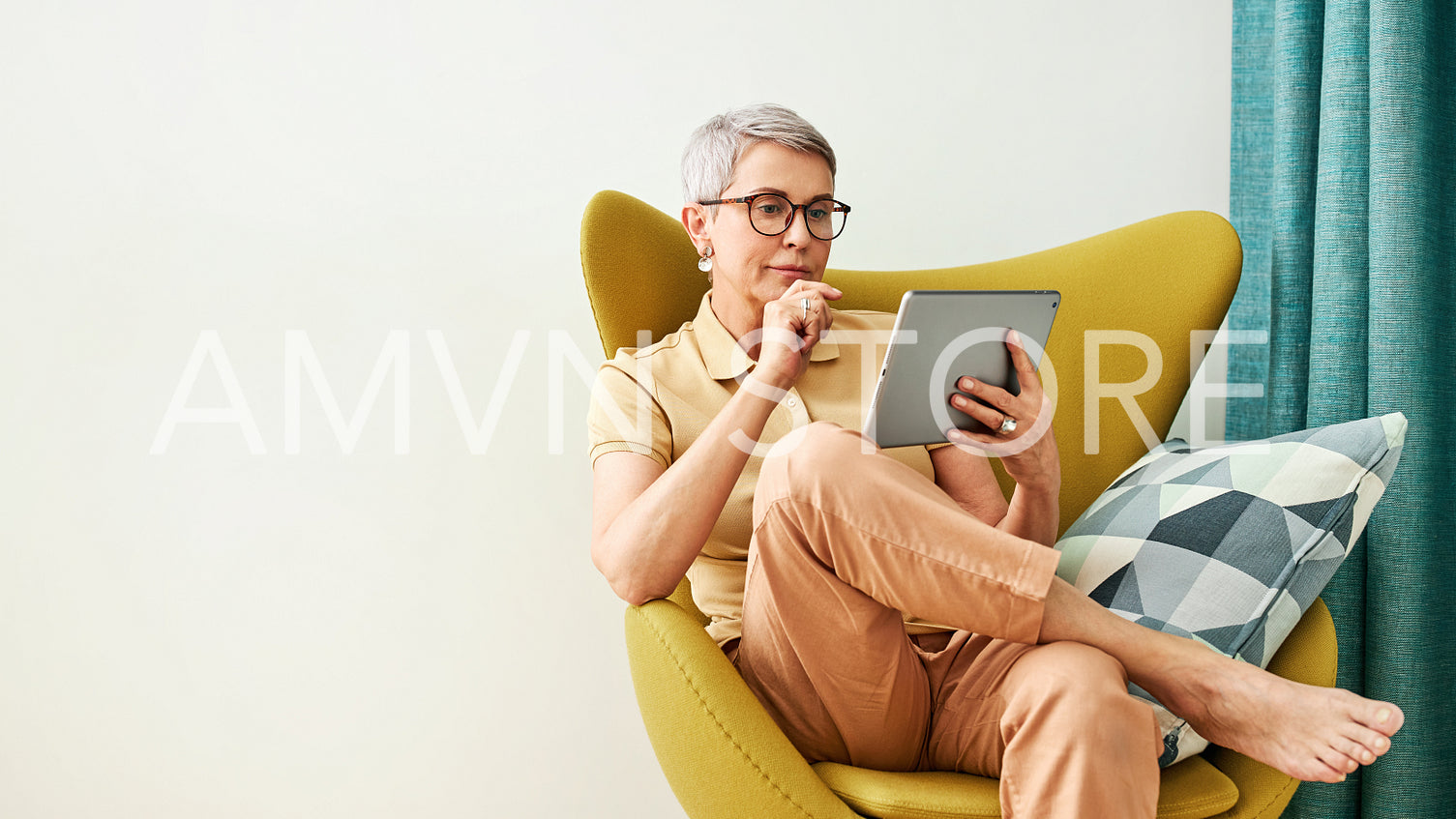 Stylish mature woman in eyeglasses thoughtfully looking at a digital tablet while sitting at home in an armchair