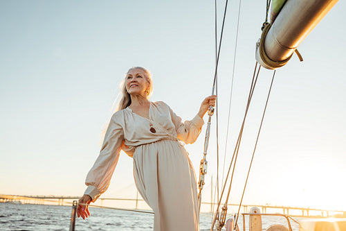 Portrait of mature woman in dress standing on yacht at sunset. Beautiful senior woman holding a rope and looking away.