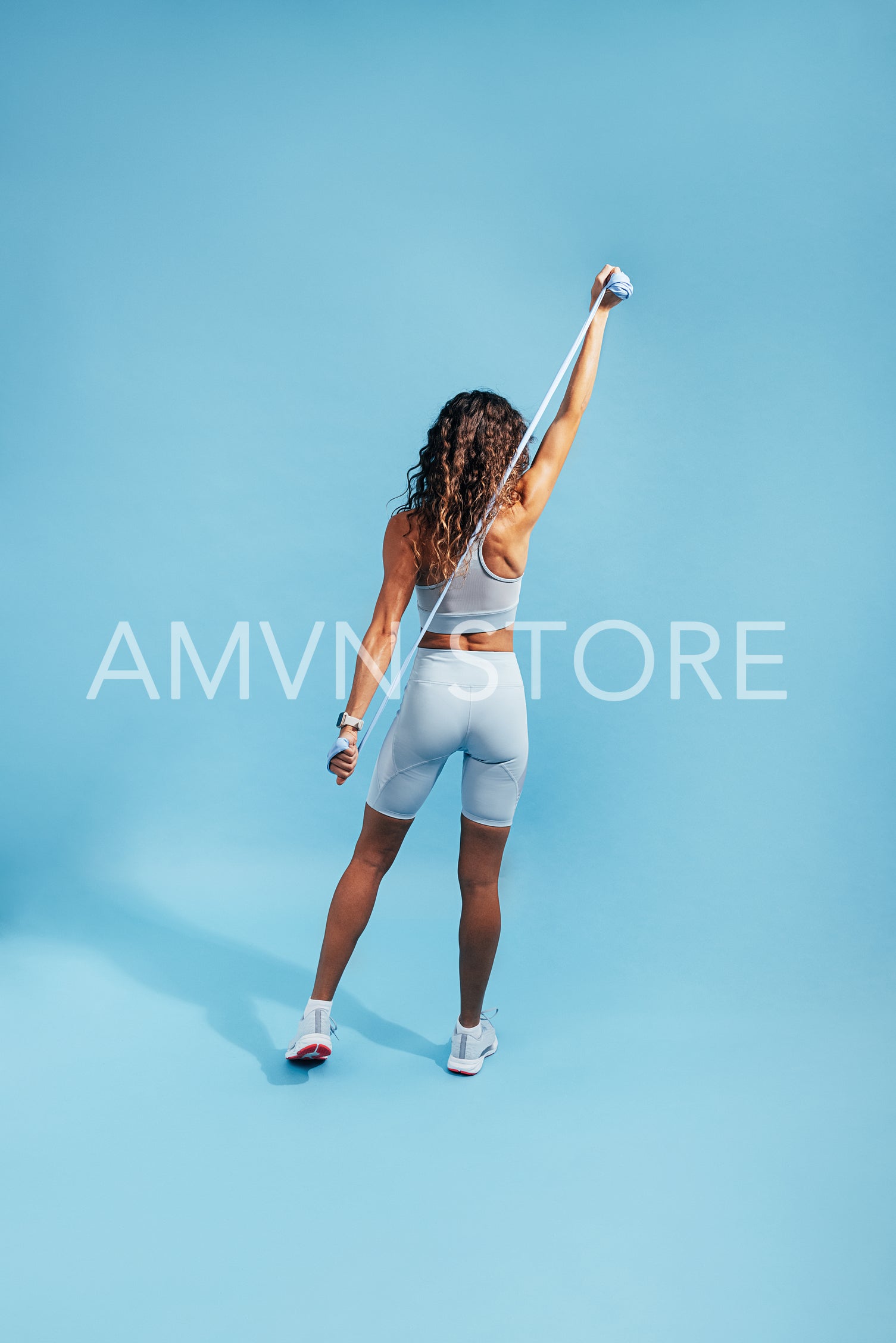 Rear view of young slim woman stretching her arms. Female warming up her hands on blue background with resistance band.