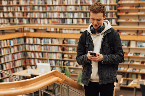 Smiling guy with a smartphone in a library. Student with headphones against bookshelves in library.